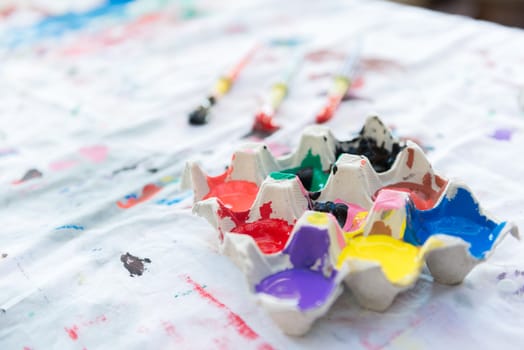 Egg carton containing color paint with paintbrushes in the background.