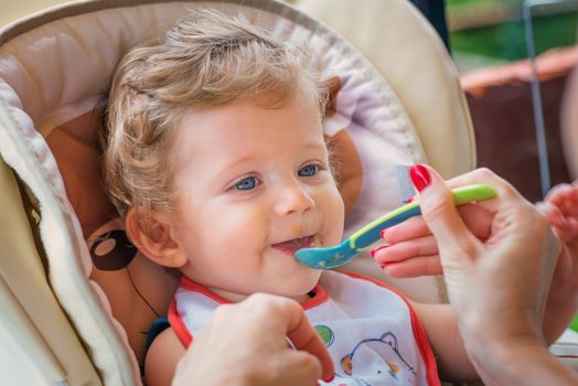 A mother feeding her baby boy with spoon outdoor.