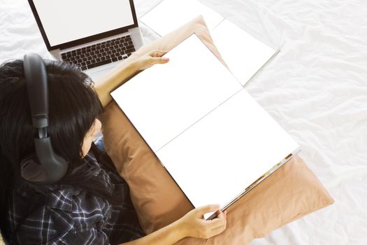 Asian girl sitting on bed reading blank book listening to music with headphone in relax mood with notebook background