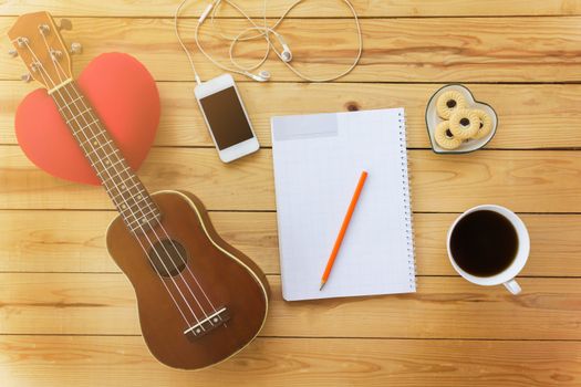 Blank note book and pencil with coffee,cookie, mobile and ukulele in vintage style on jointed wooden background