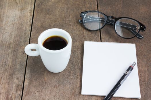 White cup of hot black coffee with blank note book and pencil on wood table with eye galsses