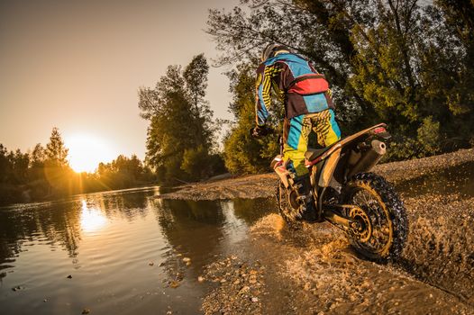 Enduro rider crossing water and muddy terrain against a beautiful sunset.