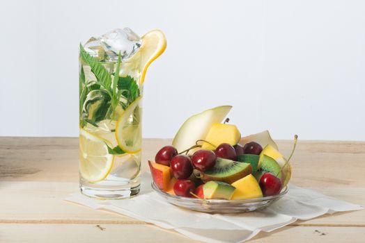 Slices of fruit, sweet cherry on a plate and lemon drink with ice in a glass on a surface from wooden boards