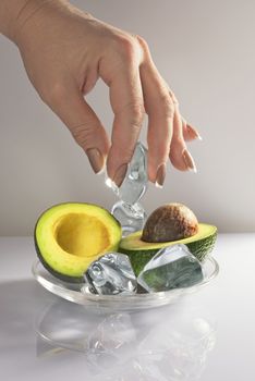 Avocado fruit cut on two parts with pieces of ice and a female hand with classical Frencha manicure