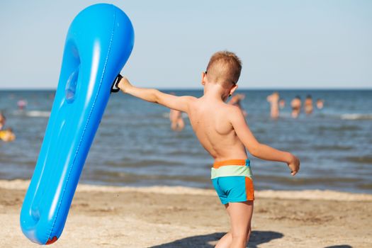 Little kid holding an inflatable mattress on the beach on hot summer day. Smiling boy playing on the beach with air mattress. Child is going to swim in the sea with swimming mattress. Summer vacation