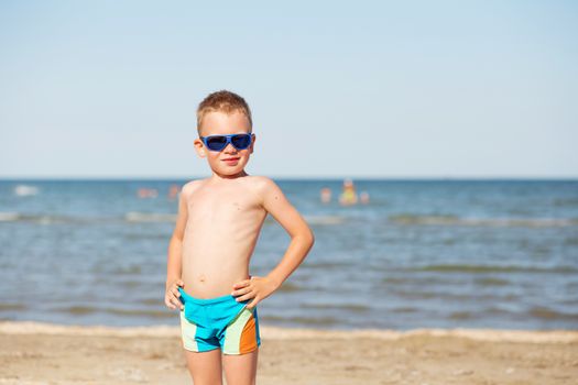 Little kid wearing fashion sunglasses on the beach on hot summer day. Smiling boy playing on the beach. Child is going to swim in the sea. Summer vacation