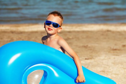 Little kid holding an inflatable mattress on the beach on hot summer day. Smiling boy playing on the beach with air mattress. Child is going to swim in the sea with swimming mattress. Summer vacation