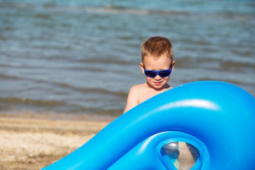Little kid holding an inflatable mattress on the beach on hot summer day. Smiling boy playing on the beach with air mattress. Child is going to swim in the sea with swimming mattress. Summer vacation