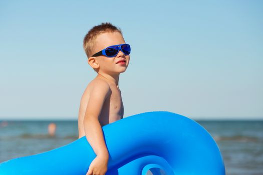 Little kid holding an inflatable mattress on the beach on hot summer day. Smiling boy playing on the beach with air mattress. Child is going to swim in the sea with swimming mattress. Summer vacation