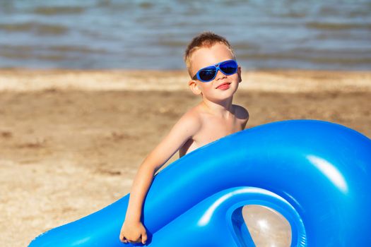 Little kid holding an inflatable mattress on the beach on hot summer day. Smiling boy playing on the beach with air mattress. Child is going to swim in the sea with swimming mattress. Summer vacation