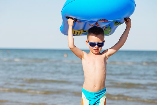 Little kid holding an inflatable mattress on the beach on hot summer day. Smiling boy playing on the beach with air mattress. Child is going to swim in the sea with swimming mattress. Summer vacation