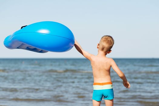 Little kid holding an inflatable mattress on the beach on hot summer day. Smiling boy playing on the beach with air mattress. Child is going to swim in the sea with swimming mattress. Summer vacation