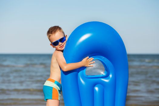 Little kid holding an inflatable mattress on the beach on hot summer day. Smiling boy playing on the beach with air mattress. Child is going to swim in the sea with swimming mattress. Summer vacation