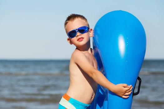 Little kid holding an inflatable mattress on the beach on hot summer day. Smiling boy playing on the beach with air mattress. Child is going to swim in the sea with swimming mattress. Summer vacation