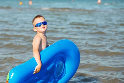 Little kid holding an inflatable mattress on the beach on hot summer day. Smiling boy playing on the beach with air mattress. Child is going to swim in the sea with swimming mattress. Summer vacation