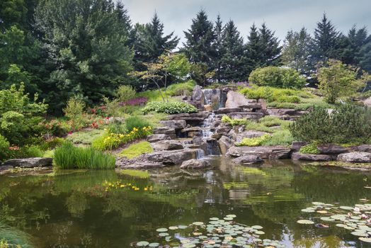 Calm water of a lake with a waterfall in a japanese garden, surrounded by trees and plants. Meijer Garden, Grand Rapids, Michigan, United States.