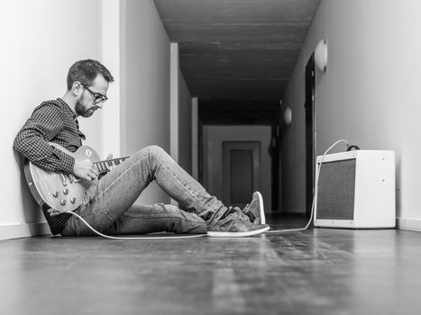 Photo of a man sitting playing his electric guitar in front of a small combo amplifier in a hallway. Black and white version.