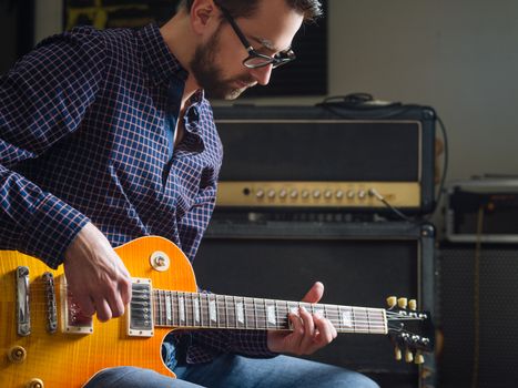 Photo of a man in his late 20's sitting in a studio playing his electric guitar.