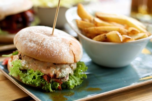 Delicious Burger with Chicken, Lettuce, Grilled Tomato and Cheese Sauce and Bowl with French Fries closeup Outdoors. Focus on Foreground