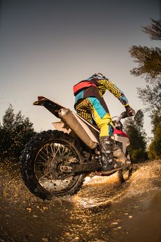 Enduro rider crossing water and muddy terrain against a beautiful sunset.