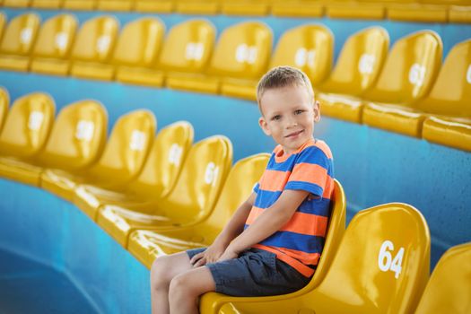 Little boy sitting in an empty stadium among yellow plastic numbered seats and applauds. Child take own seat in the stadium or dolphinarium and waiting patiently when the show or game starts.