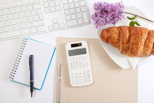 Top view office workplace - croissant, keyboard, calculator, pen, flower and notebook on white table