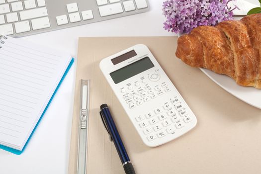 Top view office workplace - croissant, keyboard, calculator, pen, flower and notebook on white table