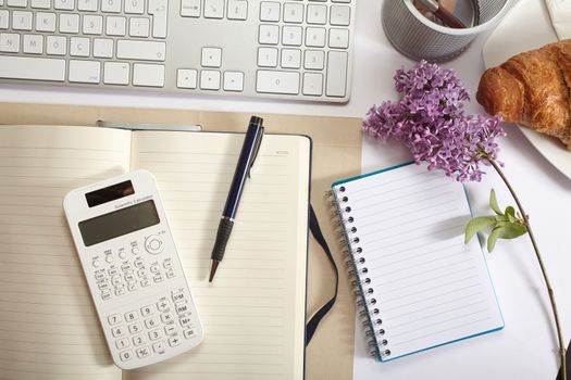 Top view office workplace - croissant, keyboard, calculator, pen, flower and notebook on white table