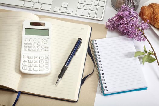 Top view office workplace - croissant, keyboard, calculator, pen, flower and notebook on white table