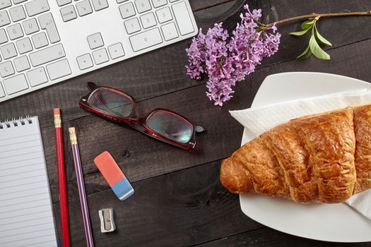 Top view office workplace - keyboard, pencil, glasses, croissant and notebook on black table