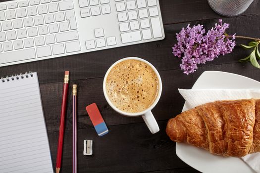 Top view office workplace - coffee, keyboard, pencil, croissant, flower and notebook on black table