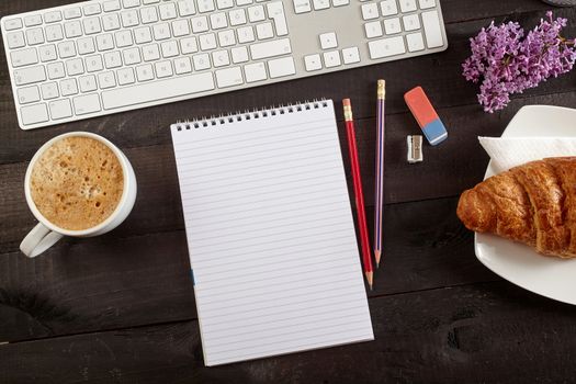 Top view office workplace - coffee, keyboard, pencil, croissant, flower and notebook on black table