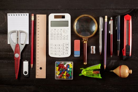 Top view office workplace - pencil, calculator, ruler, magnifying glass, pen, cutter, scissors, whirligig and notebook on black table