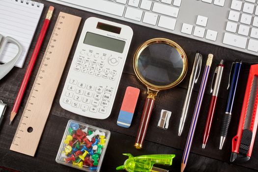 Top view office workplace - keyboard, pencil, calculator, ruler, magnifying glass, pen, cutter, scissors, whirligig and notebook on black table