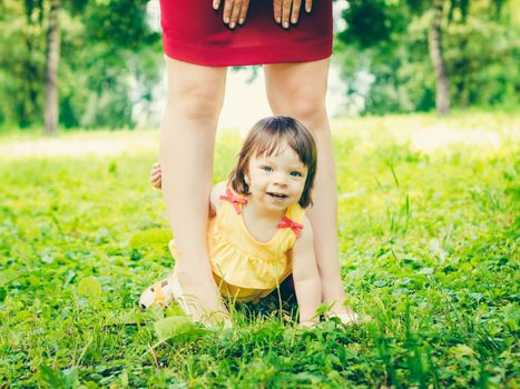 one-year old baby girl sitting between mother legs smiling and looking at camera outdoors. Daughter hiding between the mothers legs on green grass in park. Colorful image