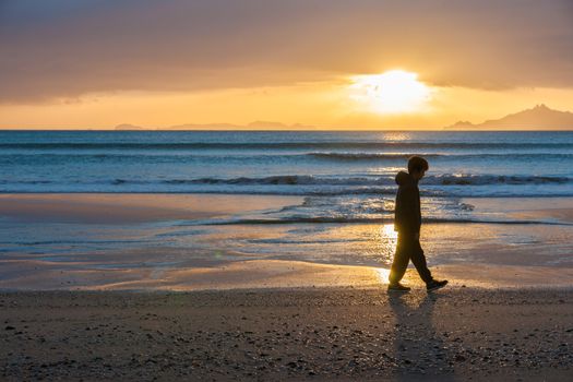 Sunrises glistening across water onto beach as a lone boy walks silhouetted at waters edge and offshore islands seen from Waipu Cove New Zealand.