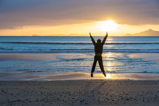 Sunrise glistens over water from horizon and off-shore islands as boy jumps with happiness on wintry morning at waters edge on beach Waipu Cove New Zealand