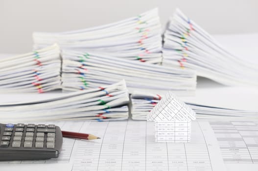 Brown pencil with calculator and house on finance account have blur pile overload paperwork of report and receipt with colorful paperclip place on white background.