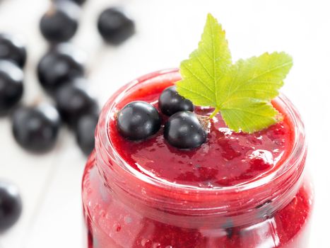 Delicious black currant smoothie or jam in glass jar with fresh berries on white wood background. Shallow DOF. Selective focus