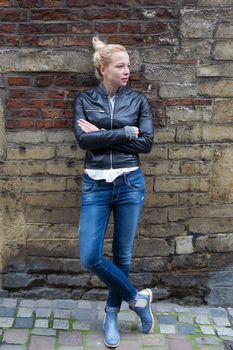 Fashion street style portrait of young beautiful calm female model, hands in pockets, leaning on vintage brick wall in background.