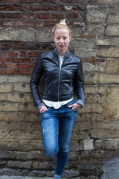 Fashion street style portrait of young beautiful calm female model, hands in pockets, leaning on vintage brick wall in background.