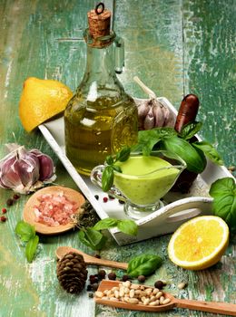 Freshly Made Creamy Pesto Sauce in Glass Gravy Boat with Raw Basil Leafs, Ingredients, Spices and Olive Oil in White Wooden Tray Arranged on Cracked Wooden background