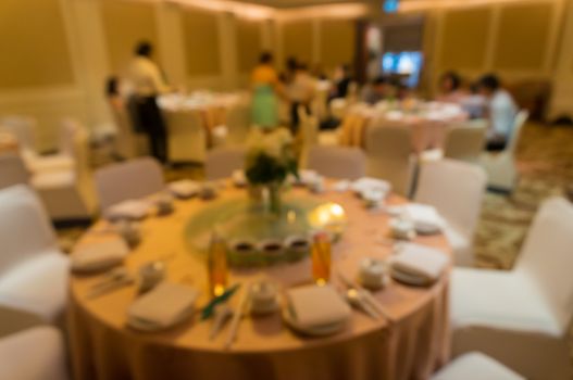 blurred image of Large dining table set for wedding, dinner or festival event with beautiful lights decoration inside large hall with people.