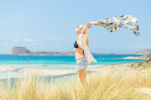 Relaxed woman, arms rised, holding colorful scarf, enjoying sun, freedom and life at beautiful beach. Young lady feeling free, relaxed and happy. Concept of vacations, freedom, joy and well being.