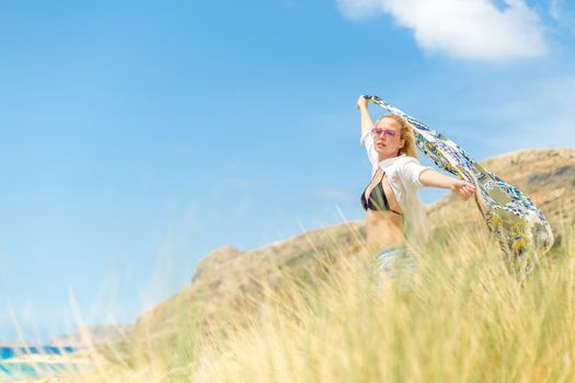 Relaxed woman, arms rised, holding colorful scarf, enjoying sun, freedom and life at beautiful beach. Young lady feeling free, relaxed and happy. Concept of vacations, freedom, joy and well being.