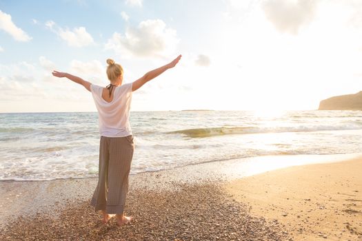 Relaxed woman, arms rised, enjoying sun, freedom and life an beautiful beach in sunset. Young lady feeling free, relaxed and happy. Concept of vacations, freedom, happiness, enjoyment and well being.