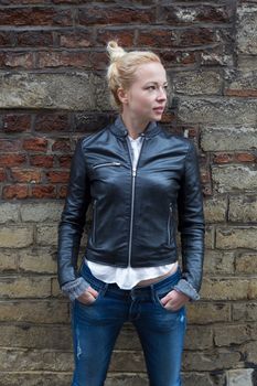 Fashion street style portrait of young beautiful calm female model, hands in pockets, leaning on vintage brick wall in background.