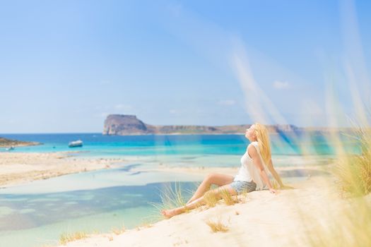 Relaxed woman enjoying sun, freedom and life an a beautiful sandy beach of Balos in Greece. Young lady feeling free, relaxed and happy. Vacations, freedom, happiness, enjoyment and well being.