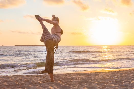 Silhouette of young woman practicing standing bow yoga pose on sandy beach at sunset.