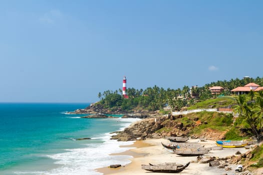 The lighthouse on the cape stretching into the Arabian Sea, beach and fishing boats at a resort in Kerala in sunny weather